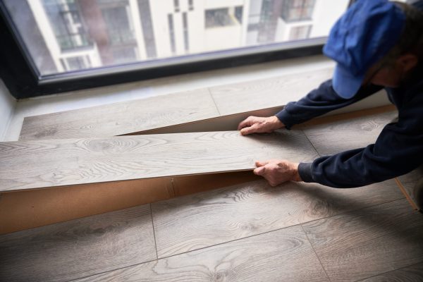 Male builder installing laminate flooring in room with large window. Man construction worker laying laminate planks on the floor in apartment under renovation. Hardwood floor renovation concept.